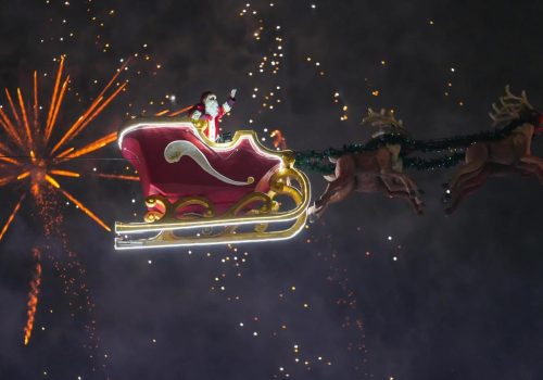 Sania Zburătoare a lui Moș Crăciun luminată pe cer, trasă de reni, în mijlocul unui spectacol de artificii de sărbători